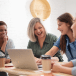 A group of four diverse women entrepreneurs laughing and looking at a laptop screen, signifying the joy of shared achievements and community support.